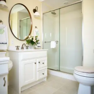 Bright bathroom with a shower, round mirror, and white vanity. Flowers add a touch of elegance.